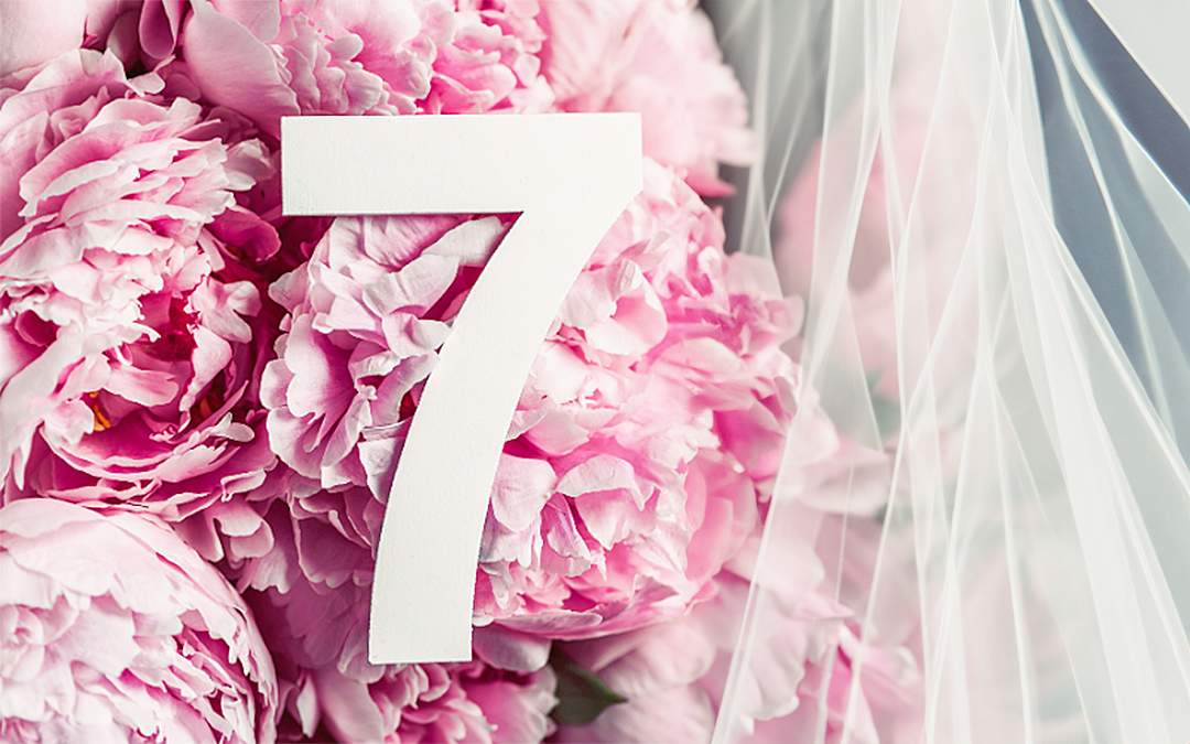 The number seven laying on top of a group of pink flowers surrounded by a white wedding veil.