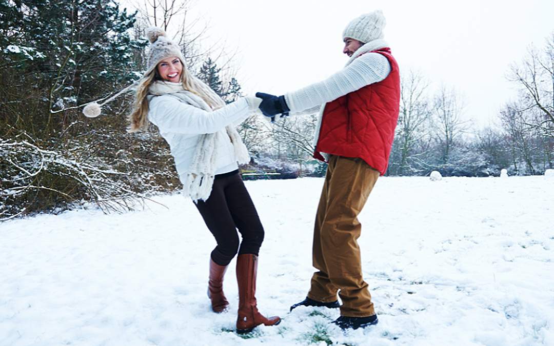 Newly engaged couple holding hands and dancing in the snow while dressed in winter clothing.