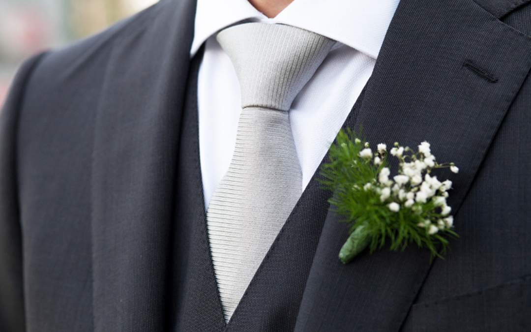 Groom wearing dark gray suit with light gray tie and white flower on lapel.