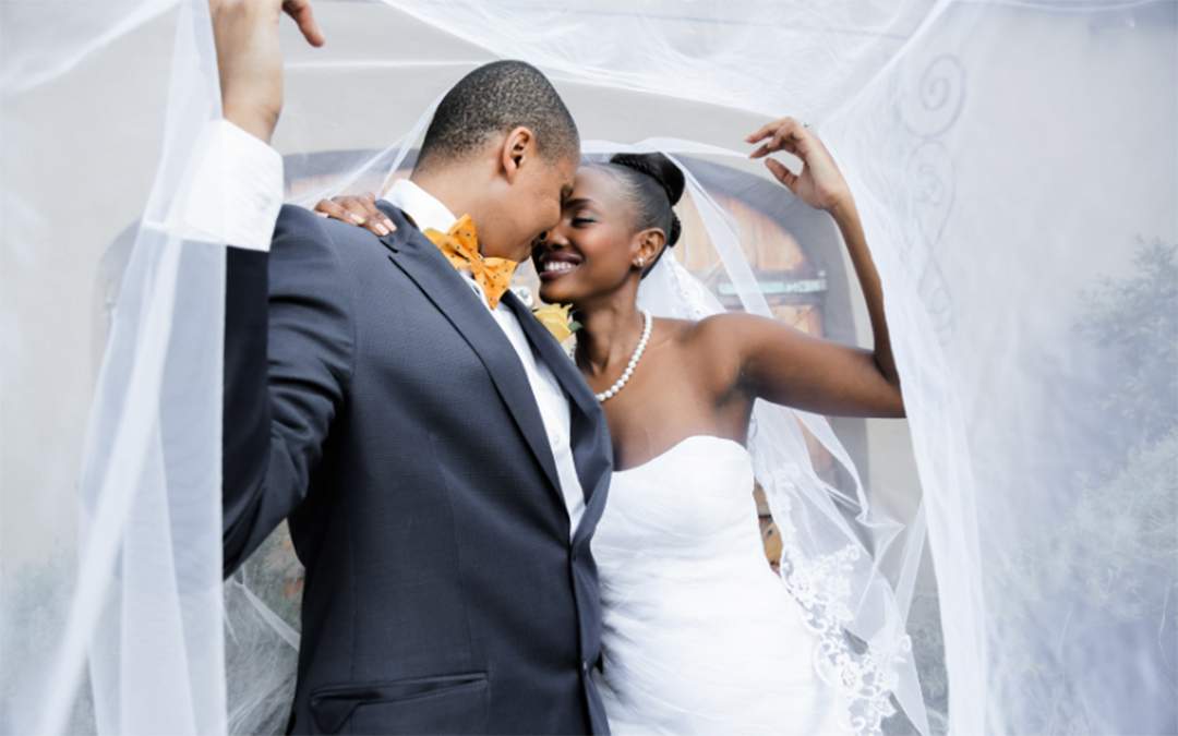 Happy bride and groom smiling at one another while standing underneath a large white veil.