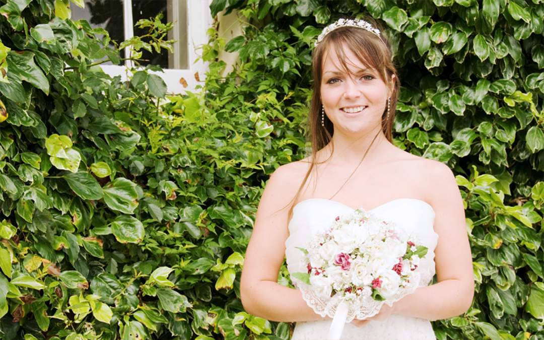 Happy bride standing in front of greenery while holding white floral bouquet.