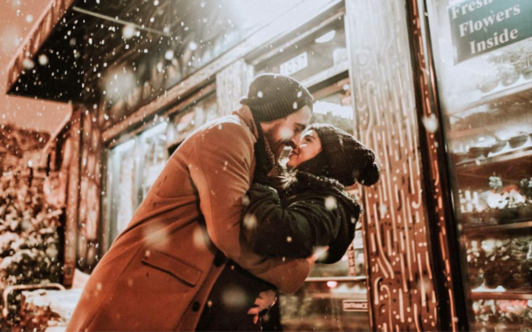 Newly engaged couple kissing in front of flower shop while it is snowing.