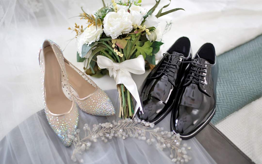 White wedding bouquet sitting between a pair of bridal shoes and a pair of grooms shoes.
