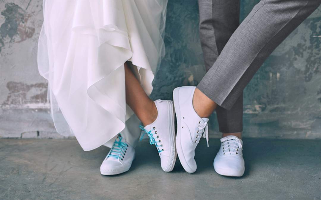 Bride and groom wearing sneakers with soles of feet touching while standing against a stone wall.